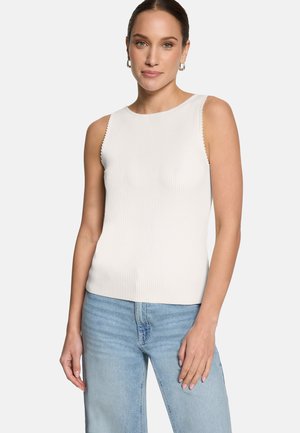 Femme vêtue d'un débardeur blanc côtelé et d'un jean bleu clair, debout avec une main reposant sur sa cuisse, sur un fond blanc.