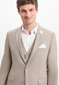 Beige strukturierte Sakkos mit einem weißen Hemd. Verfügt über einen Drei-Knopf-Verschluss, angedeutete Revers und ein weißes Taschentuch mit einer Akzentnadel.
