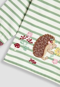 Csíkos, hosszú ujjú ing fehér és zöld színben, hímzett sünivel, virágokkal és gombákkal. Jellemzője a piros "Jojo" márkázás.