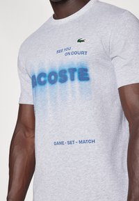 Grå bomullströja med blå textdesign, inklusive "LACOSTE" och "GAME - SET - MATCH", med en grön krokodil-logotyp som accent.