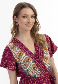 Rochie florală în fucsia vibrant, cu modele florale albe și galbene, mâneci scurte cu volane și detaliu legat la talie. Cercei circulari.