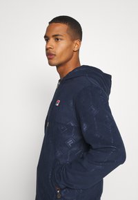 Felpa con cappuccio blu navy dotata di un design con zip, texture morbida, motivo a logo ripetuto e cappuccio. Include tasche laterali con finiture dorate.