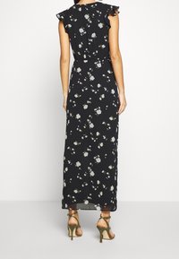 Žena v černých květinových maxi šatech bez rukávů s volánovými rameny a olivově zelených sandálech na vysokém podpatku, otočená zády.