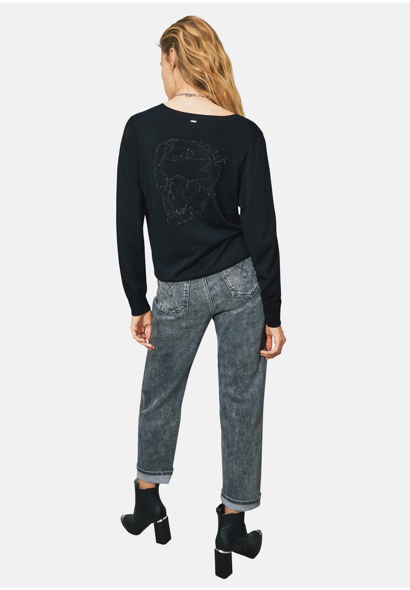 Maglione nero con design di teschio realizzato con piccoli borchietti sulla schiena. Abbinato a jeans grigi a gamba stretta e stivaletti neri.