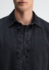 Donkerdenim overhemd met een knoopsluiting, een spreidkraag en subtiele textuur. Klein logo aan de linkerkant.