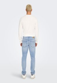 Camisa ligera blanca de manga larga combinada con jeans azul claro que cuentan con dos bolsillos traseros y zapatillas blancas, vista desde atrás.