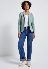 Blazer vert clair, haut blanc et jean large bleu. Le blazer possède un col cranté et un bouton unique. Porté avec des baskets blanches.