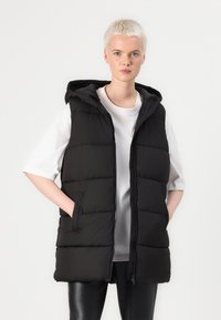 Schwarze gepolsterte Weste mit Kapuze, ausgestattet mit einem Reißverschluss vorne, seitlichen Taschen und einer glatten Textur, übereinander getragen mit einem weißen Hemd.