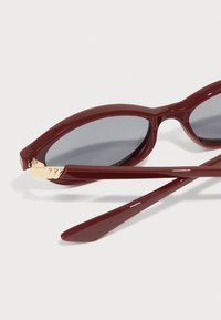 Burgundské sluneční brýle s oválnými čočkami, lesklým povrchem a zlatým akcentem na stranici. Štíhlé, rovné nožičky dotvářejí design.