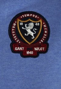 Broderad patch på ett blått tyg, med en skölddesign som har ett lejon, året "1949," och texten "GANT" och "N.H.CT" i vita och röda accenter.