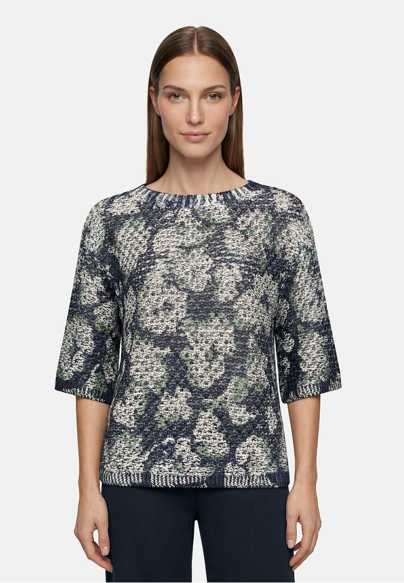 Femme portant un pull en maille texturée avec un motif abstrait multicolore et des manches jusqu’au coude, debout devant un fond uni.