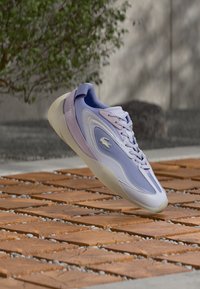 Ténis Lacoste em roxo claro e lavanda, com parte superior em malha texturada, logótipo subtil e design de sola lisa e arredondada.