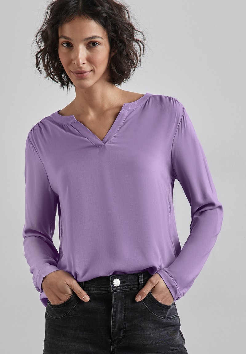 Blusa de manga larga color lavanda con escote en V y hombros fruncidos, combinada con jeans de denim oscuros. Textura suave, diseño casual.