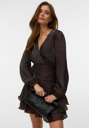 Kvinne med langt brunt hår, iført en mørk, gjennomsiktig omslagskjole med langermet, holder en svart skinnclutch mot en hvit bakgrunn.