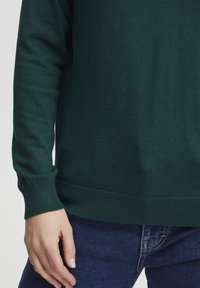 Dunkelgrüner Strickpullover mit Rundhalsausschnitt, gerippten Bündchen und Saum. Hat eine weiche Textur. Kombiniert mit blauen Denim-Jeans.
