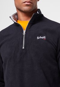 Pullover in pile nero con mezza cerniera, interno del colletto grigio e logo "Schott NYC" ricamato in bianco sul petto. Tessuto morbido.