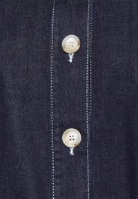 Tumma denimpaita, jossa on kaksi suurta, pyöreää valkoista nappia, näkyvät saumat ja lähikuva napituslistan tekstuurista.