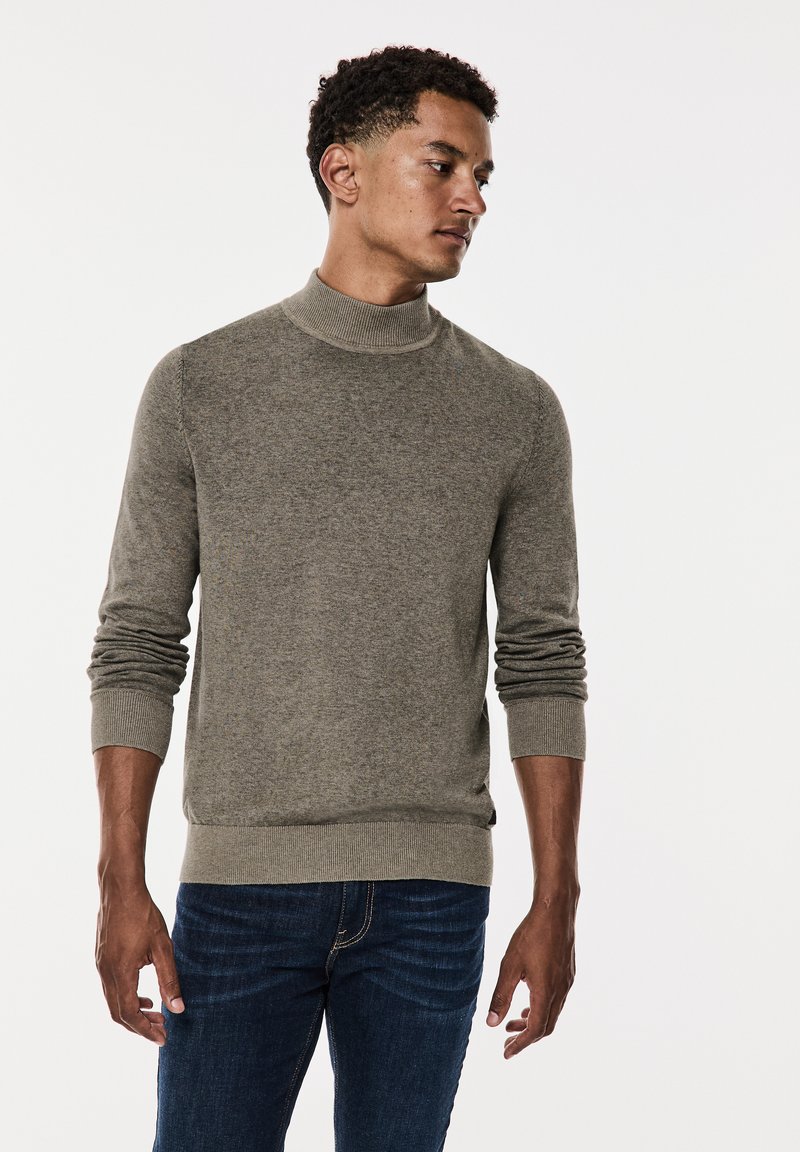 Pull à col roulé en kaki en tissu doux, avec des poignets et un ourlet côtelés, porté avec un jean en denim foncé. Design simple et ajusté.