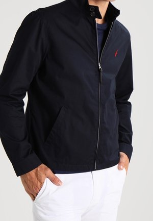 Mann trägt eine dunkelblaue Jacke mit Reißverschluss und kleinem rotem Logo, darunter ein dunkelblaues Hemd und weiße Hose, Hände in den Taschen.