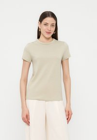 Γυναίκα με ίσια σκούρα μαλλιά που φορά χρυσούς κρίκους, μπεζ κοντομάνικο πουκάμισο και λευκό φαρδύ παντελόνι σε απλό λευκό φόντο.