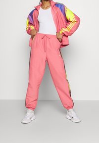 Rosa träningsdräkt med kontrasterande gula och blå detaljer, bestående av en zipjacka och byxor med dragsko. Vita sneakers kompletterar looken.