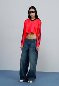 Rotes, kurzes Sweatshirt mit schwarzen Streifen an den Ärmeln, kombiniert mit lockeren blauen Jeans und weißen Slippern. Das Modell trägt eine Sonnenbrille.