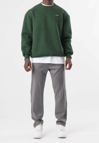 Uomo che indossa una felpa verde scuro con il testo "MILLION", una camicia bianca sotto, pantaloni grigi con righe nere ai lati e sneaker bianche su uno sfondo bianco.