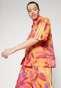 Cămașă cu mânecă scurtă, de culoare portocalie, cu modele vibrante de frunze tropicale în nuanțe de roz și violet; are trei dungi verticale galbene pe mâneci.