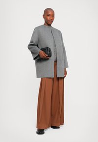 Femeie îmbrăcată într-o haină gri, pantaloni largi maro și pantofi negri, ținând un clutch negru matlasat pe un fundal alb simplu.