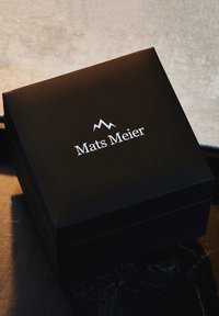Boîte rectangulaire noire avec une finition mate, présentant un logo argenté "Mats Meier" et un graphique de montagne sur le couvercle.