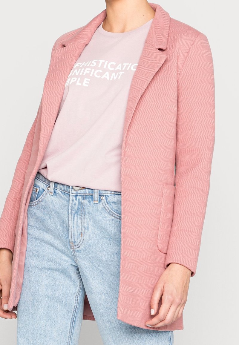 Manteau texturé rose clair avec un revers cranté et deux poches avant, porté sur un T-shirt graphique rose pâle et un jean bleu clair.