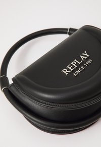 Sac à main en simili cuir noir avec une forme incurvée et une poignée circulaire. Il arbore un logo argenté portant la mention "REPLAY SINCE 1981." Texture lisse.