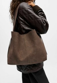 Sac en toile en daim marron avec une texture douce, ouverture sur le dessus et sans quincaillerie visible, porté contre une veste matelassée marron.