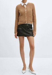 Brązowy sweter z dzianiny z guzikami, dwoma przednimi kieszeniami, w zestawieniu z czarną mini spódnicą z zamkami błyskawicznymi oraz białymi czółenkami na spiczastym obcasie.