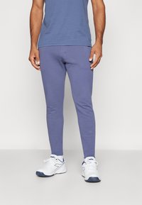 Eng anliegende lila Jogginghosen aus weichem Material, mit glatter Textur und minimalistischem Design, kombiniert mit weißen Sportschuhen und Socken.