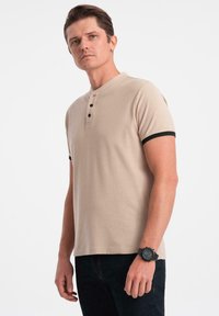 Polo shirt in cotone beige con maniche corte, rifinitura nera e colletto a tre bottoni. Indossato con jeans scuri e un orologio nero.