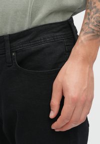Vaqueros de mezclilla negros con corte slim y una textura suave. Tienen un bolsillo delantero y un cierre de botón visible. Mano descansando sobre el vaquero.