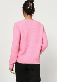 Pull rose en tissu tricot doux, avec un col rond et des manches longues, doté de poignets et d'un ourlet côtelés. Porté avec un pantalon noir.