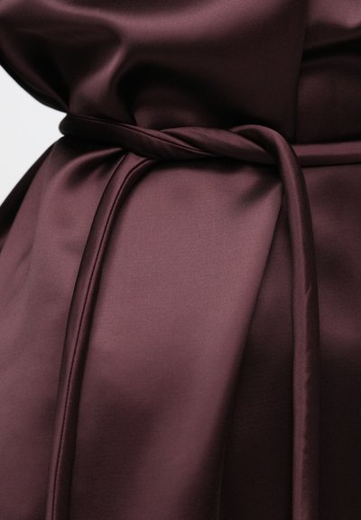 Λαμπερό ύφασμα σε βαθύ βουργουνδί χρώμα, μαζεμένο και δεμένο σε έναν απλό κόμπο με μια λεπτή ζώνη στην μέση που ταιριάζει στο χρώμα.