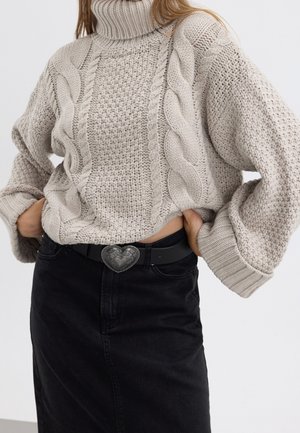 Kvinde iført beige chunky kabelstrikket højhalset sweater med ballonærmer, stoppet ind i sort nederdel med høj talje og hjerteformet bæltespænde.