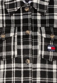 Černobílá kostkovaná flanelová košile s límečkem, dvěma náprsními kapsami a stříbrnými knoflíky; obsahuje vyšívané logo "TOMMY JEANS" v barvách.