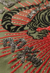 Lona verde con un tigre bordado en negro y rayos de costura roja, con texto negro de cuentas y acentos metálicos.