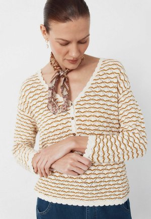 Kobieta związaną z tyłu włosami, ubrana w brązowy, wzorzysty szalik na szyję oraz kremowy sweter z brązowymi falistymi pasami, spoglądająca w dół z założonymi złożonymi rękami.