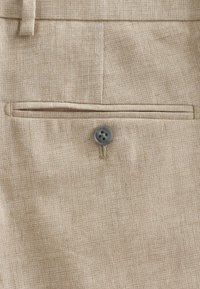 Tela texturizada en color beige con un bolsillo trasero que presenta un botón gris. La tela tiene un sutil patrón de tejido sin brillo visible.