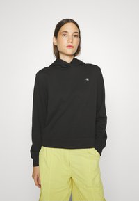 Czarny bawełniany hoodie z przednią kieszenią i logo na klatce piersiowej, w zestawie z żółtymi spodniami cargo. Posiada sznurki w kapturze i ściągacze na mankietach.