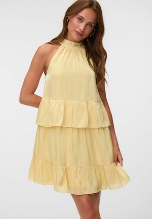 Femme aux longs cheveux bruns portant une robe sans manches jaune clair à étages avec un col montant, se tenant devant un fond clair uni.