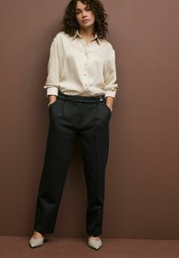 Cremfarbene Bluse mit langen Ärmeln, kombiniert mit schwarzen Nadelstreifen-Hosen. Das Model trägt spitze beige High Heels und hat die Hände in den Taschen.