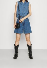 Denim ärmelloses Hemd mit geknoteter Vorderseite, kombiniert mit blauen Denim-Shorts und schwarzen Cowboy-Stiefeln. Eine kleine schwarze Umhängetasche wird getragen.