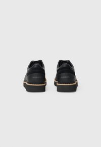 Ein Paar schwarze SOREL-Schuhe mit dicken schwarzen Sohlen und beige Nähten, von hinten auf einem weißen Hintergrund gezeigt.