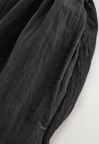 Vêtement en tissu noir avec une texture froissée, présentant des coutures visibles et un design décontracté et fluide. Les bords sont légèrement effilochés.